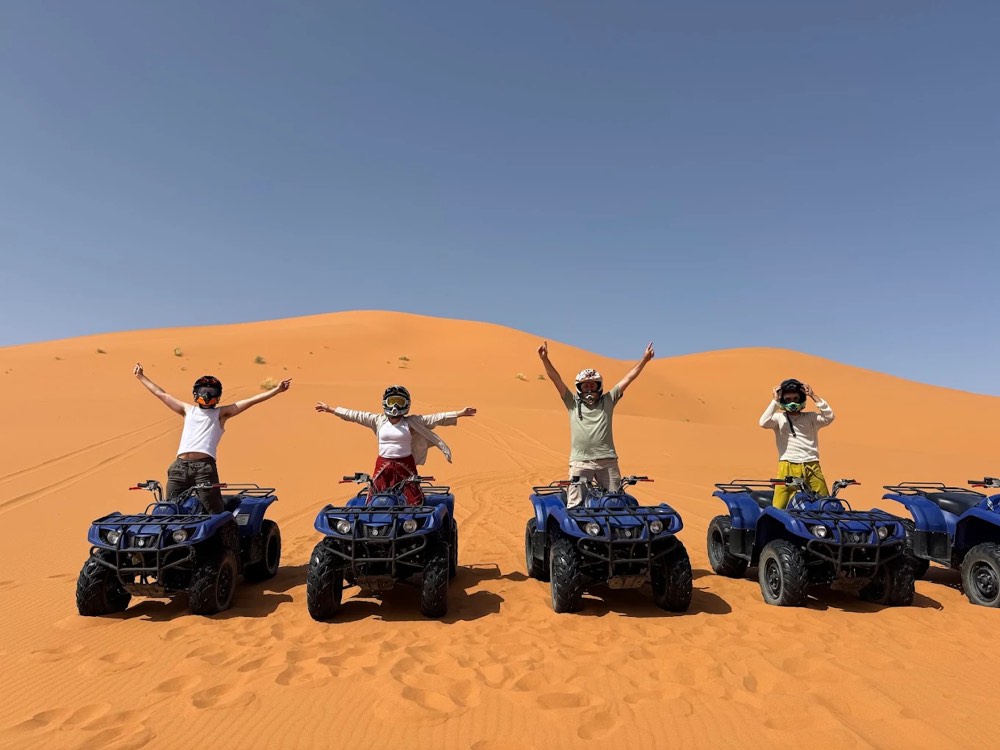 Quattro piloti su quad blu alzano le braccia su una duna dell’Erg Chebbi