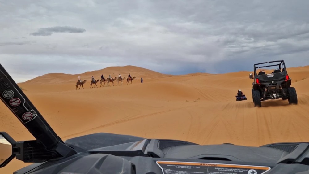 View from inside a buggy towards another buggy and a camel caravan in the dunes